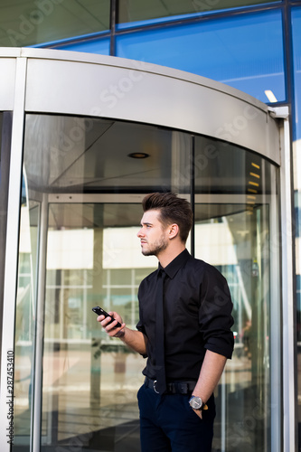 Man professional office worker waiting call on mobile phone while standing outdoors near company against glass door during break. Male intelligent lawyer using cellphone standing outside enterprise