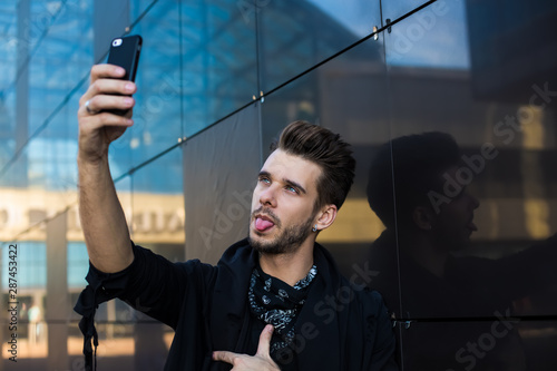 Hipster guy showing tongue while shooting video on mobile phone, standing outdoors near modern building. Handsome man photographing hisself on cellphone front camera, having fun photo