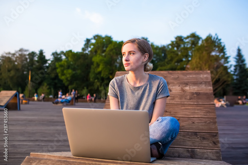 Thoughtful woman enjoying view after online webinar on modern laptop computer, sitting on wooden bench in park in sunny summer evening. Pondering female dreaming after work on netbook device