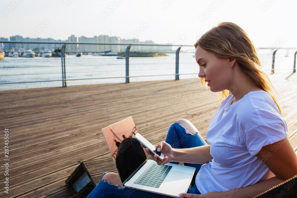Attractive woman using applications on mobile phone after training course on laptop computer while sitting on sea promenade in sunny summer evening, Female chatting in social network via cellphone