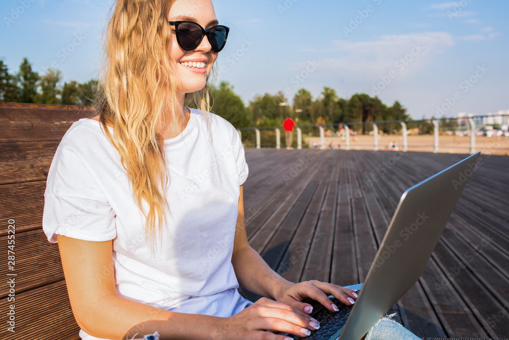 Happy smiling hipster girl with stylish look having distance work in internet via laptop computer while siting outdoors in sunny summer day. Female publication specialist typing on notebook