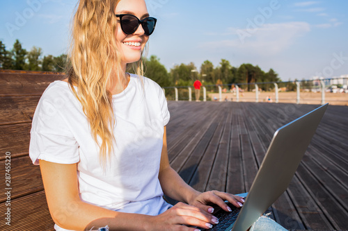Happy smiling hipster girl with stylish look having distance work in internet via laptop computer while siting outdoors in sunny summer day. Female publication specialist typing on notebook