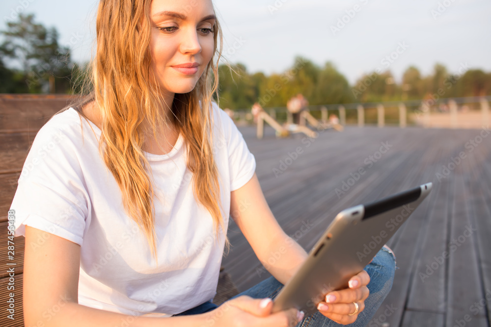 Young beautiful blonde woman reading e-book on modern touch pad while relaxing in the fresh air in sunny summer day during leisure. Charming Caucasian female watching video on digital gadget