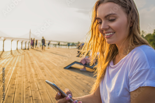 Happy smiling hipster girl watching video in social network via smartphone while sitting outdoors near copy space for promotional content. Cheerful female sending sms via cellphone during walking time