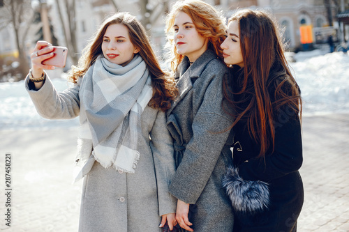 three beautiful elegant girls in winter coats walking together in a sunny winter city and use the phone © hetmanstock2