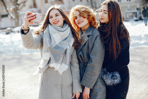 three beautiful elegant girls in winter coats walking together in a sunny winter city and use the phone