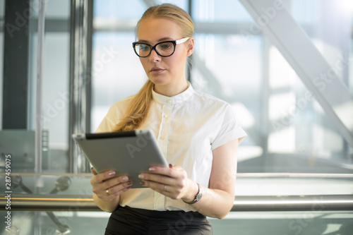 Serious woman in eye glasses dressed in office wear smart secretary reading business news on web site via touch pad gadget while standing inside enterprise. Female prosperous banker using applications