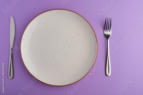Empty white dish with knife and fork on a purple background, with copy space for your menu or recipe. Menu card for restaurants and table setting.Photo Flat lay