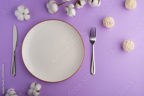 Empty white dish with knife and fork on a purple table background with a cotton branch, with copy space for your menu or recipe. Menu card for restaurants and table setting. Horizontal photo. Flat lay