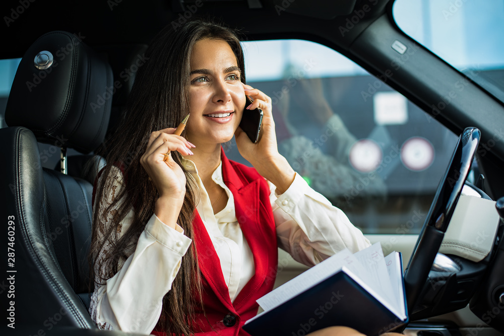 Happy smiling woman successful business owner having pleasant mobile phone conversation and using notepad for notary, sitting in luxury automobile. Joyful female office worker talking via cellphone