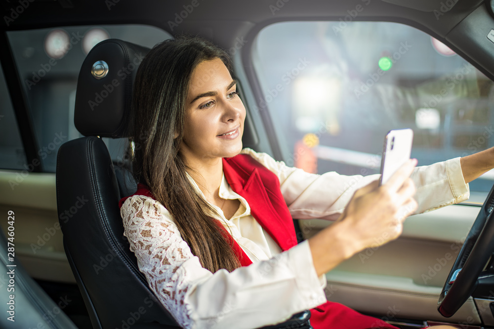 Happy smiling woman dressed in luxury formal wear reading pleasant text message on mobile phone while sitting behind the wheel of a modern car. Cheerful female using applications on cell telephone