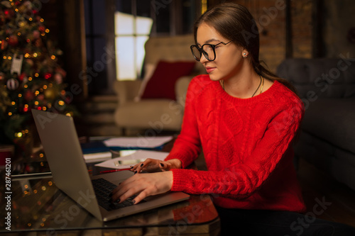 Hipster girl in stylish glasses professional fashion magazine editor checking e-mail on laptop computer while sitting in home interior near Christmas tree during holidays in winter. Online learning