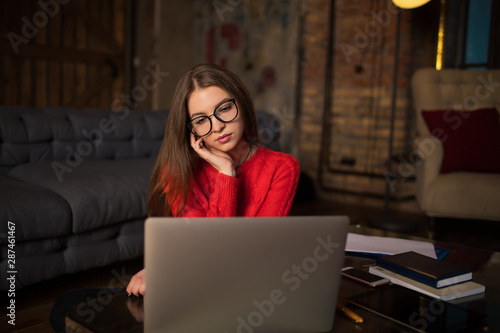 Young hipster girl in stylish spectacles watching movie via pc laptop computer, sitting in home. Female having webinar via netbook during learning. Woman reading news via notebook