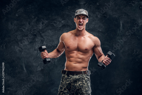 Attractive shirtless man in cap is doing exercises with dumbbells and screaming.