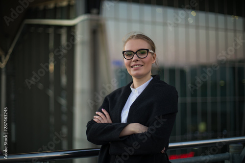 Portrait of cheerful confident woman leadership in stylish glasses and formal wear standing with crossed arms outdoors against company building. Female successful executive director smiling for camera
