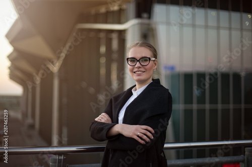 Portrait of a happy smiling woman successful business worker in fashionable spectacles and autumn coat standing with crossed arms outside company after work day. Female CEO looking in camera