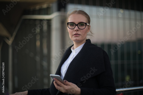 Portrait of a serious confident woman professional banker in fashionable spectacles and autumn coat holding in hand mobile phone and looking in camera while standing against modern building