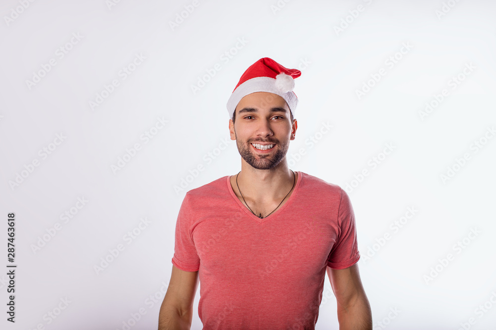Portrait of a happy smiling bearded man in casual pink shirt and Canta Clause hat looking in camera while standing isolated in studio against white background with copy space for promotional content