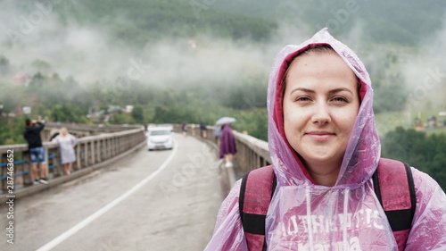 Close-up of a traveler girl smiling in a sweatshirt, straps, raincoat, with drops on a bridge with people and cars, mountains with trees, in Montenegro