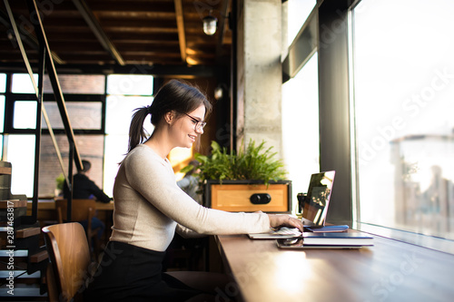 Smiling woman clever university student having online learning via pc laptop computer while sitting in co-working space. Cheerful fashionable hipster girl in spectacles having webinar via notebook