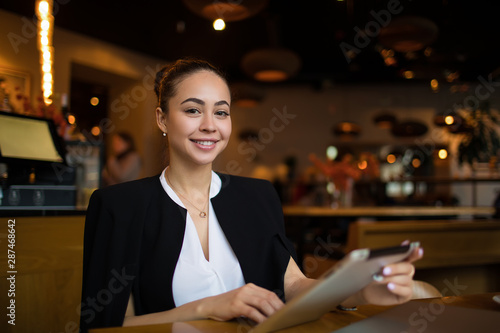 Portrait of a happy smiling woman professional hostess holding touch pad in hands and looking in camera, sitting in restaurant interior. Beautiful female business owner using digital tablet
