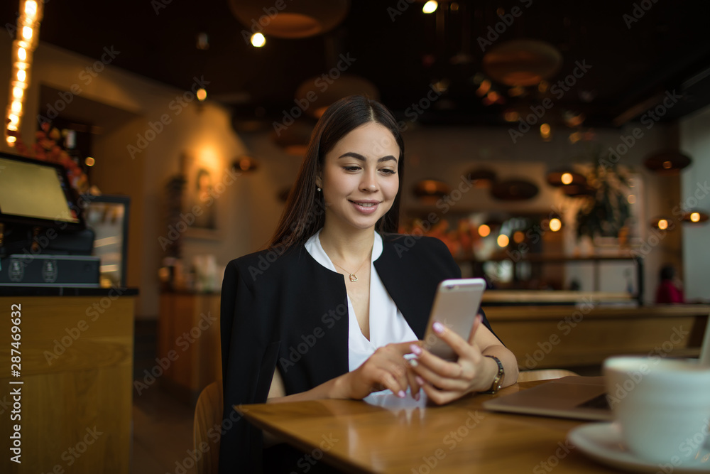 Charming woman in luxury wear checks the status of the account on mobile phone while resting in coffee shop during free time. Smiling pretty female manager online chatting on cellphone