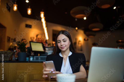 Attractive woman real estate analyst reading e-mail on mobil phone while sitting in restaurant during coffee break. Female leadership browsing wifi on smartphone gadget  resting in cafe during leisure