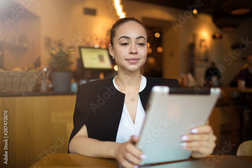 Young attractive woman clever university student online learning via portable touch pad computer while resting in restaurant. Female reading e-book via digital tablet, sitting in coffee shop