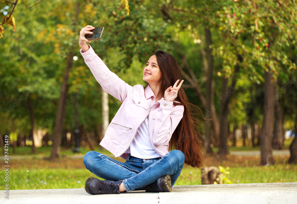 Brunette girl photographs herself on a cell phone while sitting in an autumn park.