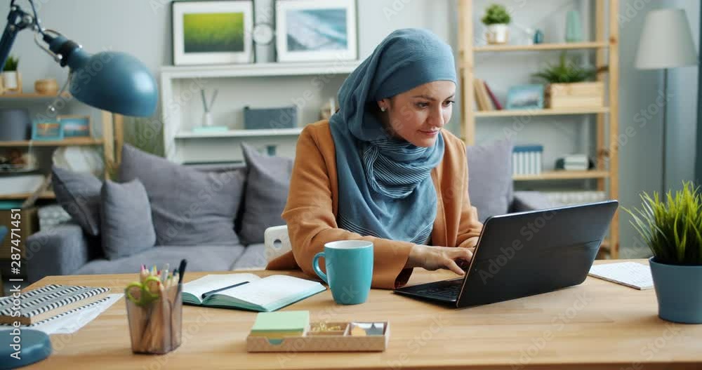 Independent girl in hijab freelancer working with laptop in apartment typing sitting at desk alone. Modern people, communication and technology concept. Independent girl in hijab freelancer working with laptop in apartment typing sitting at desk alone. Modern people, communication and technology concept.