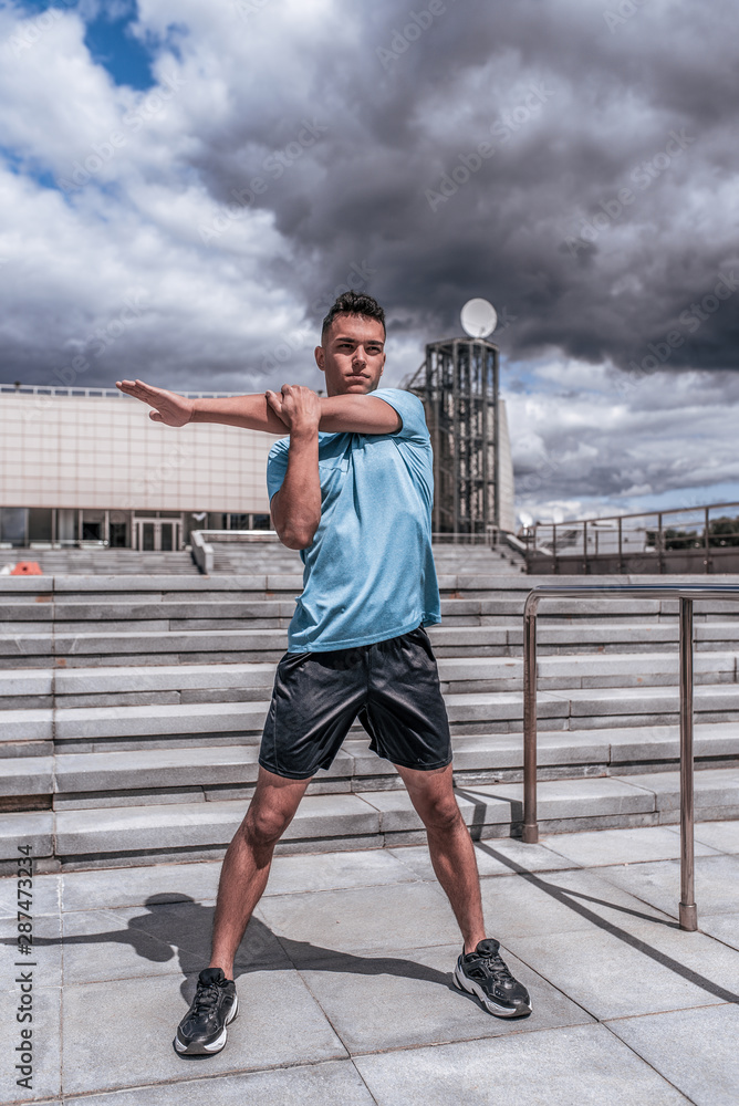 Fototapeta premium Athlete man doing shoulder muscle stretch before training. In the summer in the city, preparation for fitness workout, sportswear shorts t-shirt and sneakers. Step cloud background.