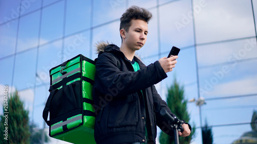 delivery man with green backpack and a scooter is looking for the client's address in the phone photo