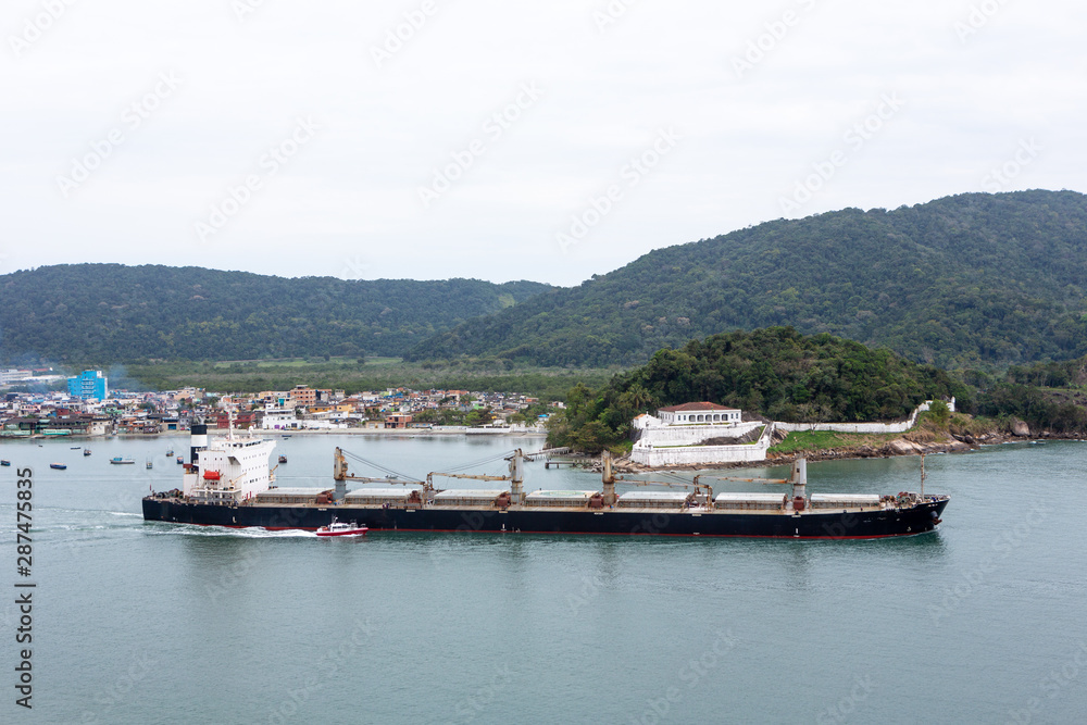 Aerial view of cargo ship sailing out of Santos port through shipping channel carrying soybeans for export. Departure with the help of harbor support boat. Logistics, shipping and commerce concept. Aerial view of cargo ship sailing out of Santos port through shipping channel carrying soybeans for export. Departure with the help of harbor support boat. Logistics, shipping and commerce concept.