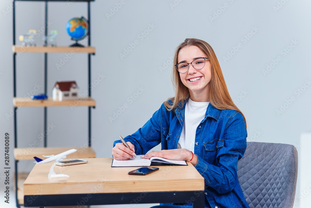 Smiling hipster girl in eyeglasses works and makes some notes and looking at camera. Smiling hipster girl in eyeglasses works and makes some notes and looking at camera.