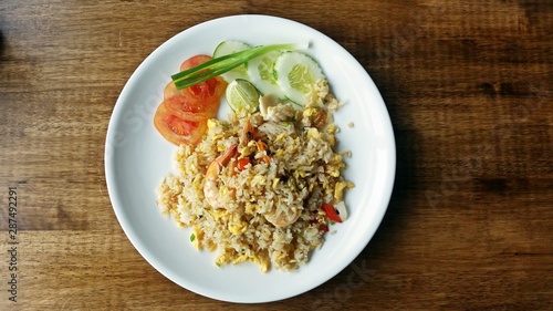 Seafood fried rice with tomato slices, cucumber slices and scallion in white plate on wooden table.