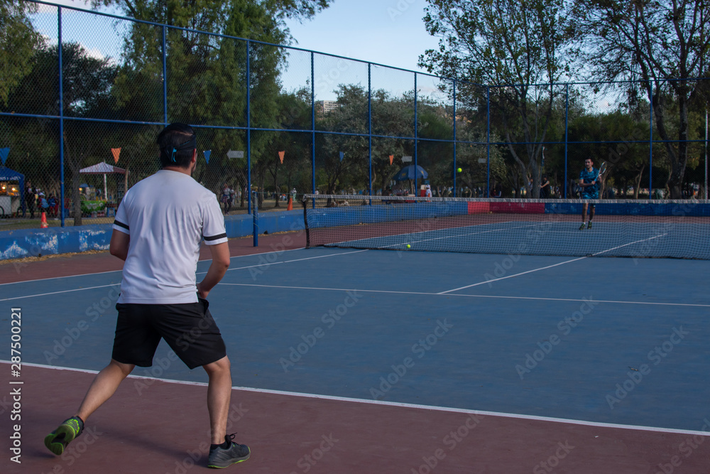 Fototapeta premium Hombre adulto latino de camiseta blanca en pantaloneta negra jugando tenis con amigo en cancha profesional hispano