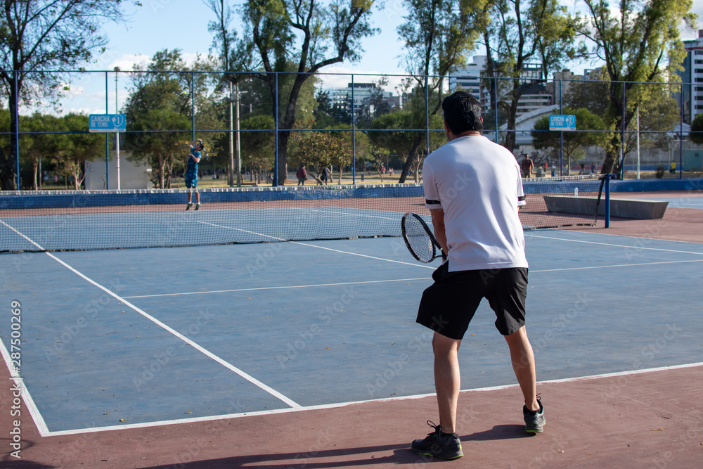 Hombre adulto latino de camiseta blanca en pantaloneta negra jugando tenis  con amigo en cancha profesional  hispano