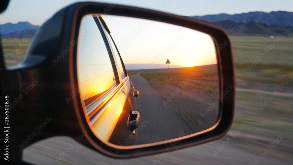 View in the side mirror of the car. Orange dawn beyond the hills. The car goes at speed. Visible green fields, grass, grasslands. Black color of the car.