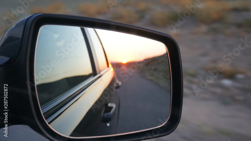 View in the side mirror of the car. Orange dawn beyond the hills. The car goes at speed. Visible green fields, grass, grasslands. Black color of the car.