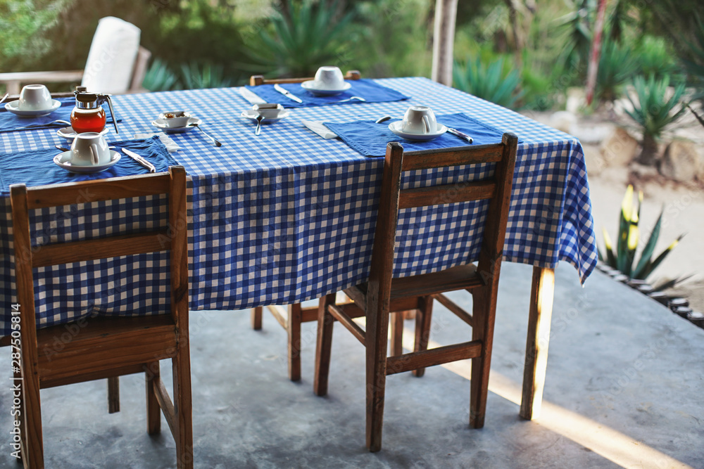 Table with empty plates and coffee cups,  blue chequered tablecloth, ready for morning breakfast, blurred green foliage in background - tropical holiday resort