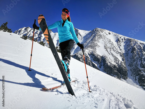 Young woman in light blue jacket standing, smiling happy with mountaineering equipment, showing one ski, sun shines on snow, bright sky over mountains behind photo