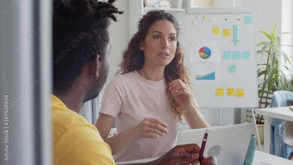 Panning of Caucasian woman with long curly hair and African man wearing casual clothes sitting at table in office and discussing important issues Panning of Caucasian woman with long curly hair and African man wearing casual clothes sitting at table in office and discussing important issues