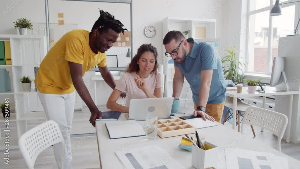 Pan shot of two diverse men and Caucasian woman with long curly hair gathered at laptop in office and having talk Pan shot of two diverse men and Caucasian woman with long curly hair gathered at laptop in office and having talk