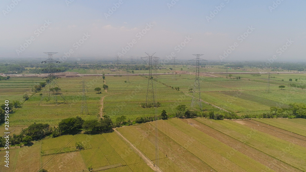 Electricity pylons bearing power supply across agricultural land with sown green, rice fields in countryside. aerial view power pylons and high voltage lines java, indonesia.High voltage metal post