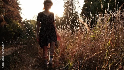 Girl walks in the high grass and spikelets toward the camera with the sun hitting her back and light coming through her. Trendy hipster girl enjoying nature, touching spikelets during sunrise photo