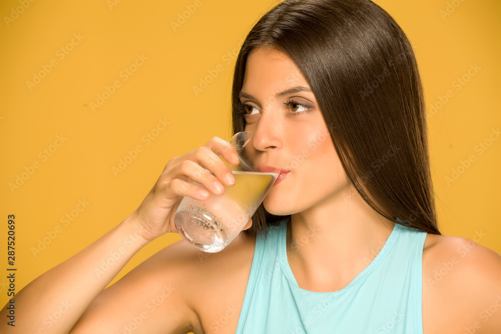 Young smiling woman drinking water from a glass on yellow background Young smiling woman drinking water from a glass on yellow background