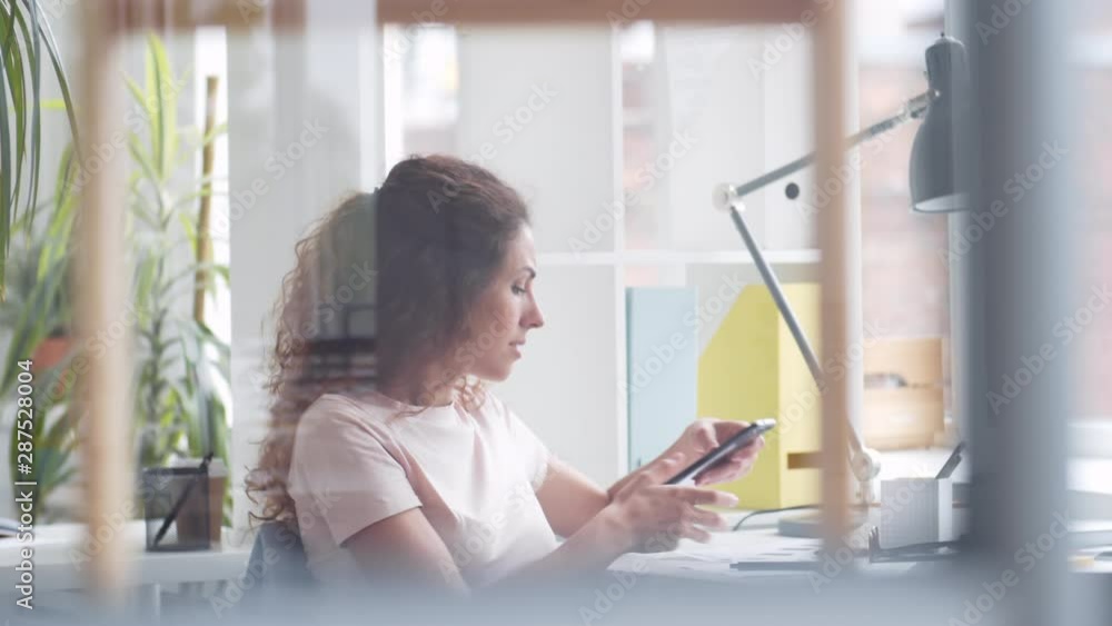Pan shot of Caucasian casual businesswoman with long curly hair sitting at desktop and having pleasant phone communication Pan shot of Caucasian casual businesswoman with long curly hair sitting at desktop and having pleasant phone communication