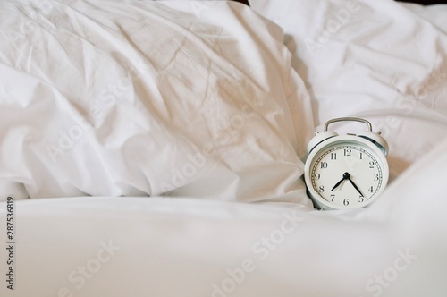 Analog vintage alarm clock is on white bed, time in the morning with a bright sunshine from the outside of window