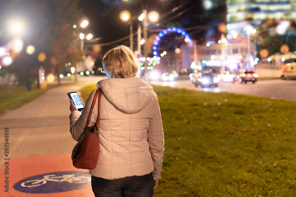 Fototapeta premium A woman with a mobile phone in her hand is walking through the night city.
