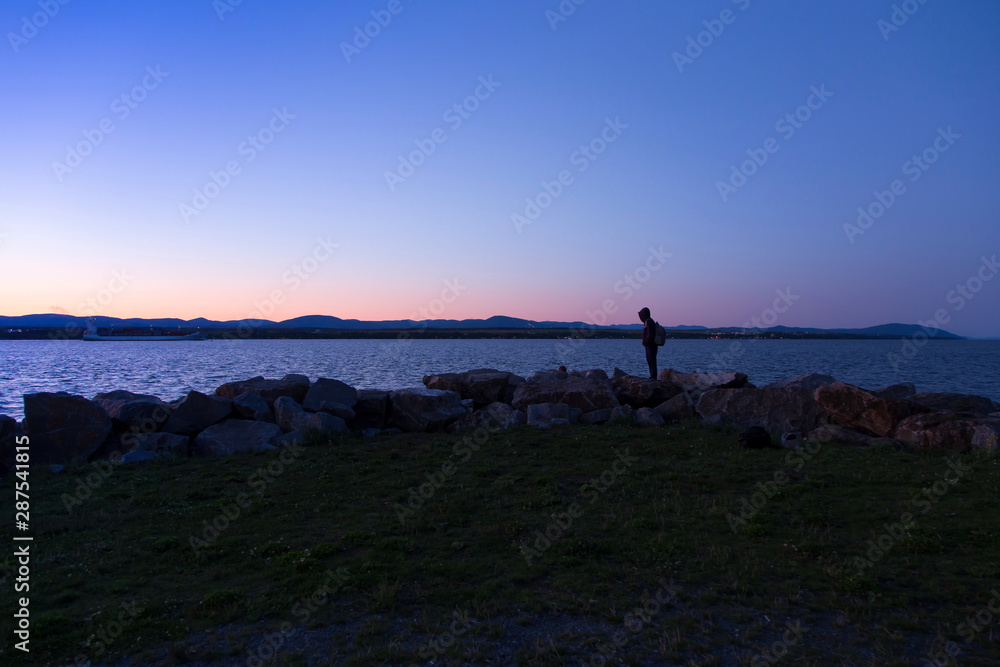Teenager standing in profile on boulders during a summer evening blue hour, with the St. Lawrence River, the Island of Orleans, and the Laurentians in the background, St-Michel-de-Bellechasse, Quebec